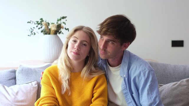 Portrait of a beautiful young heterosexual couple sitting on a cozy couch, looking at the camera and cuddling affectionately in their bright and modern living room at home
