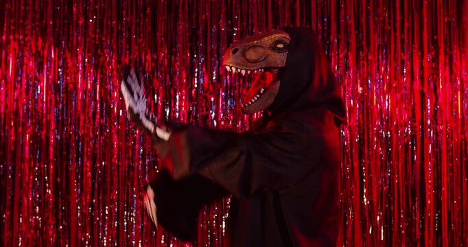 Halloween dance and cosplay of person in funny dinosaur mask and costume in front of tinsel curtains. Dragon character wearing black cloak and skeleton gloves, dancing with circle motion of arms