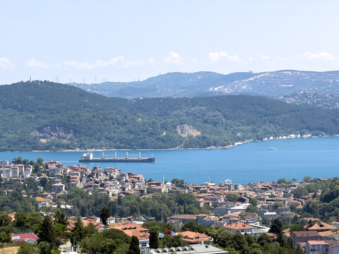 View of a cargo ship sailing through the Bosphorus strait, with dense urban landscape, terracotta rooftops, and lush green hills, Istanbul, Istanbul, Turkiye.