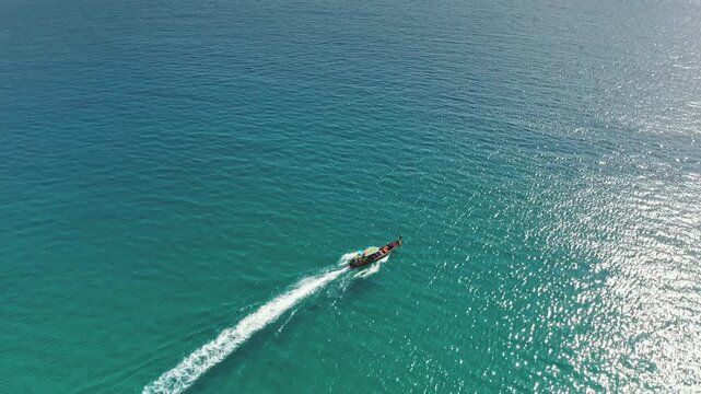 Longtail boat creating a white wake travels across vibrant turquoise waters of the Andaman Sea, showcasing tropical travel and summer vacation concepts in Phuket, Thailand. Aerial view drone flight