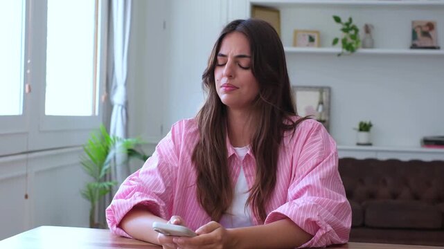 Upset young woman sitting at a table in her living room, reacting with shock and sadness after reading a bad text message on her smartphone, feeling heartbroken and disappointed by the news