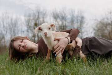Obraz premium Happy woman lying on grass outdoors, cuddling a goat kid, showcasing a warm lifestyle moment, affectionate bond with animals, serene countryside vibe, relaxed smile