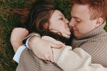 Obraz premium Smiling couple lying in the grass, embracing in a cozy sweater, enjoying a peaceful outdoor moment. Warm natural light highlights affection, comfort, and casual romance and connection.