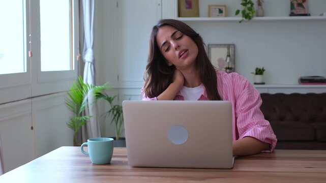 Exhausted young woman suffering from neck pain and stiffness while working on her laptop at home, feeling tired from her sedentary job and bad posture while massaging her sore muscles