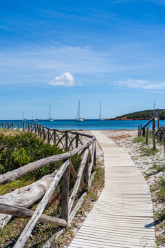 View of the wooden boardwalk leading to the turquoise waters of Spiaggia I due Mari with sailboats anchored in the bay in La Maddalena, Sardegna, Italy.