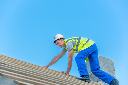 Building a house, a worker is carefully climbing the wooden roof framework, wearing a hard hat and a high visibility vest under a clear blue sky, indicating outdoor construction work and safety