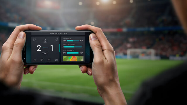 Person holding a mobile phone displaying live soccer match statistics with score updates during a professional game in a stadium