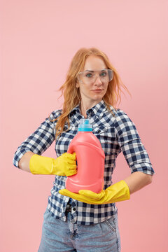 Young woman confidently holding a pink detergent bottle and wearing protective yellow gloves with safety glasses, ready for household chores, on a solid pink background