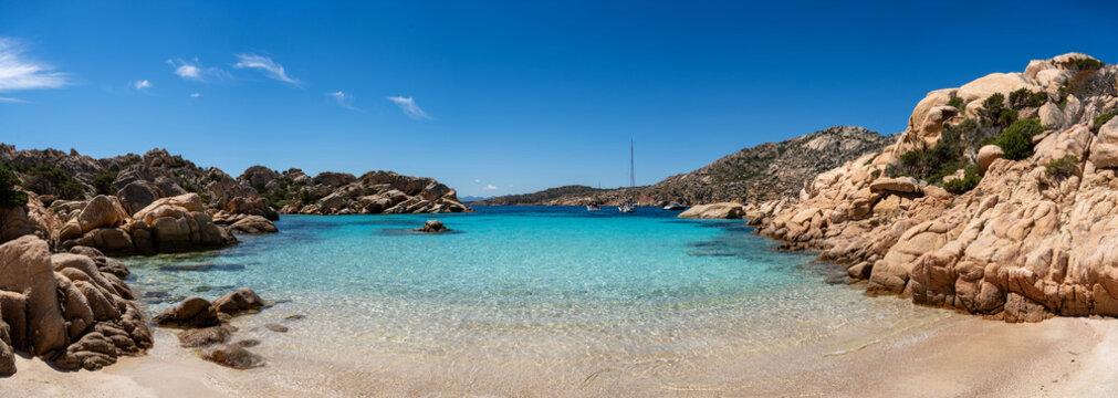 View of Cala Coticcio Beach with turquoise water, granite rock formations, and sailboats in the background in La Maddalena, Sassari, Italy.