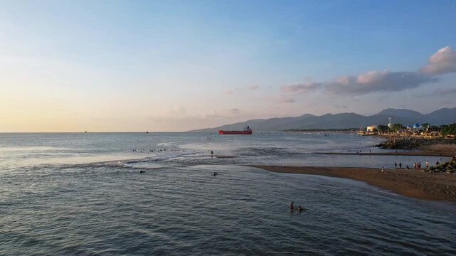 Aerial drone footage of Mataram city beach front in Lombok island, Indonesia, with silhouettes of young local surfers on little waves, a tanker boat behind, at late sunset, beachfront buildings