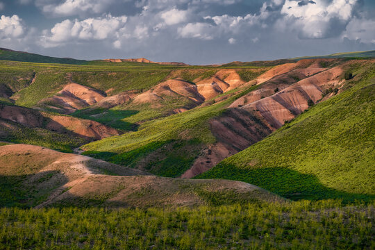 View of rolling hills and rugged terrain near Arashan Lake, with green vegetation and reddish-brown soil under a cloudy sky in Pop District, Namangan Region, Uzbekistan.