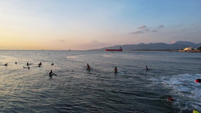 Aerial drone footage of Mataram city beach front in Lombok island, Indonesia, with silhouettes of young local surfers on little waves, a tanker boat behind, at late sunset, with mountains behind