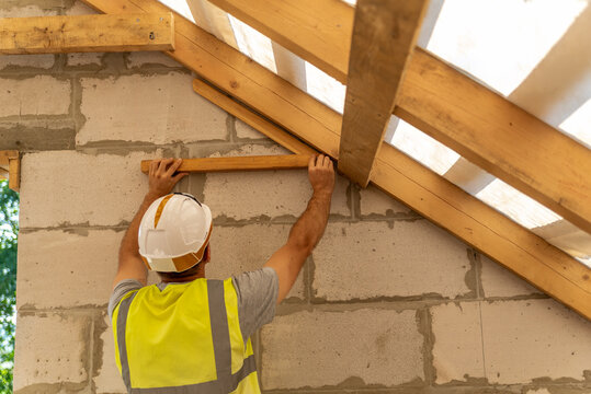 Construction worker wearing a hard hat and safety vest, installing wooden beams for a roof frame on a new residential house building site, focusing on structural elements