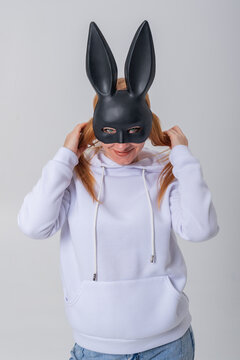 Young redhead woman in a black rabbit mask and white hoodie playfully pulling her pigtails, smiling with a mischievous, mysterious vibe in a clean studio portrait on white background