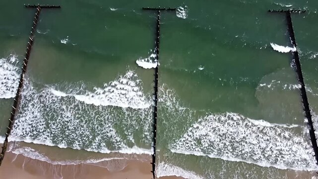 Drone view of waves between wooden groynes on beach.