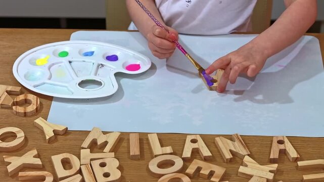 Creative child doing arts and crafts, coloring wooden alphabet blocks with acrylic paint. Letter A. Close-up of kid's hands painting a wooden letter blue, craft activity at home. Little child painting