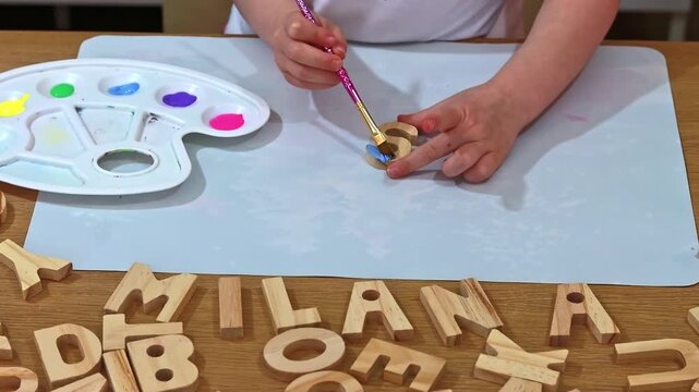 Creative child doing arts and crafts, coloring wooden alphabet blocks with acrylic paint. Letter S. Close-up of kid's hands painting a wooden letter blue, craft activity at home. Little child painting