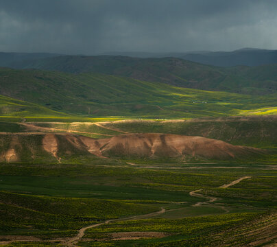 View of Arashan Lake surrounded by rolling green hills, dirt paths, and dramatic stormy skies in Pop District, Namangan Region, Uzbekistan.