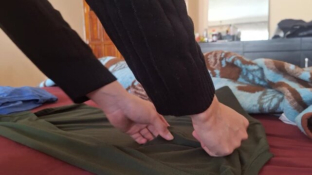 Hands fold a green shirt on a bed. Warm tones suggest a calm, home environment.