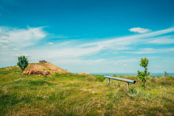 Obraz premium Peaceful rural hilltop landscape featuring tall grass, scattered trees and a rusted metal pipe beneath a vast blue sky