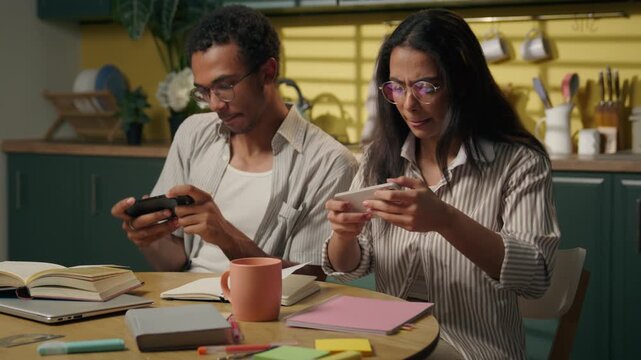 Focused African American man and Latina woman playing mobile games at home multiracial students friends using mobiles studying online gaming gadgets gen z teenagers esports screen addiction at kitchen
