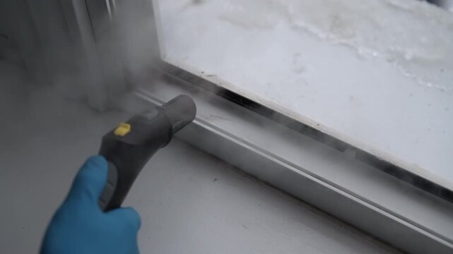 Close-up hand of cleaning service employee in blue glove using professional steam cleaner to sanitize dirty white plastic window frame, then wiping away grime and moisture with green microfiber cloth.