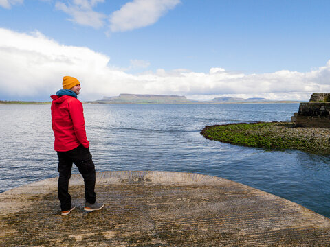 Person in red jacket and yellow hat stands on stone pier. Sea stretches wide under dramatic cloudy sky. Dark storm clouds loom over water horizon. County Sligo, Ireland. Benbulbin mountain viewpoint.