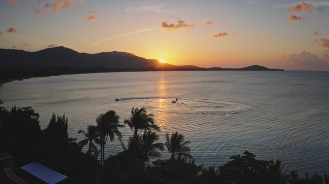 Golden hour sun setting over the calm ocean illuminating jet skis playing on the water with palm tree silhouettes and mountains in the background creating a beautiful Thailand travel scene in Samui