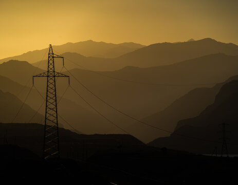 View of an electricity pylon silhouetted against the layered mountain ranges of the Arashan Lake area during golden hour in Pop District, Namangan Region, Uzbekistan.