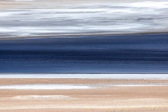 View of the Salar de Aguas Calientes salt flat with white salt crust, deep blue water, and tan mineral sediment in the arid landscape of San Pedro de Atacama, Antofagasta, Chile.