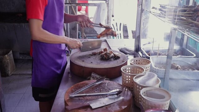 Man Seller Cuts & Chops Fresh Grilled BBQ Thai Style Chicken (Gai Yang) on a Tamarind Cutting Board. Isaan Asian Street Food Restaurant in Bangkok Thailand 