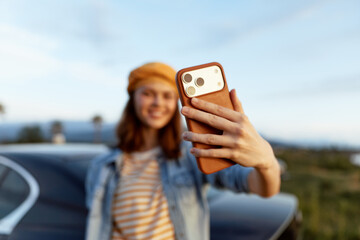 Obraz premium Cheerful woman outdoors, taking a selfie, smiling with sunlit glow, casual denim jacket, roadside scenery, warm tones, relaxed lifestyle, bright day