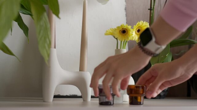 Hand places vase with yellow flowers on shelf beside candles and green plants. Concept of home decor and room decoration