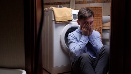 Fototapeta premium Distressed man feeling desperate, covering his face with hands, sitting alone on the floor in a laundry room, experiencing mental health issues and extreme sadness