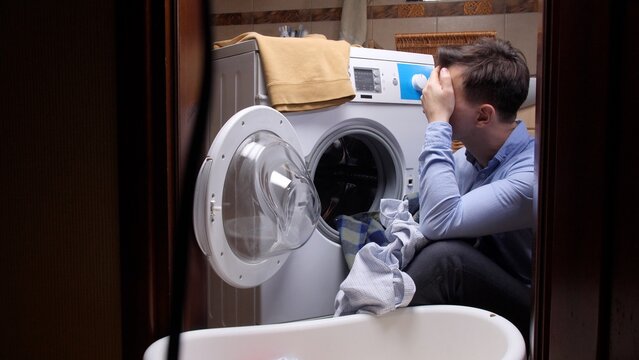 Frustrated man sitting on the floor in front of an open washing machine door with dirty laundry, covering his face with his hand, symbolizing appliance breakdown and household stress