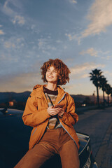 Fototapeta premium Woman lifestyle sunset outdoors portrait, smiling with curly hair, orange jacket, sitting by car on street, holding strap, warm evening light, palm trees softly blurred.