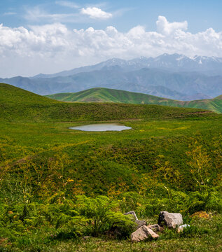 View of Arashan ko'li surrounded by lush green rolling hills and distant snow-capped mountains under a bright sky in Pop District, Namangan Region, Uzbekistan.