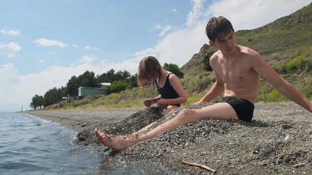 Handsome teenage boy and girl sitting on seashore with feet in water and covering their feet with pebbles. Pebble beach instead of sand. Wild beach with rocky coastline. Brother and sister