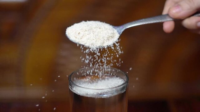 Pouring Psyllium Husk Powder From Spoon Into Glass of Water Close Up. Adding Isabgol Dietary Fiber Supplement to Glass for Digestion Health. Isabgol in Water for Gut Wellness with Healthy Drink.