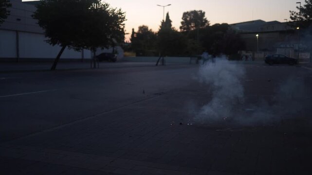 Smoking firecracker exploding and sparking on an empty city street at dusk, creating a bright flash and sparks while people watch from a safe distance in the background