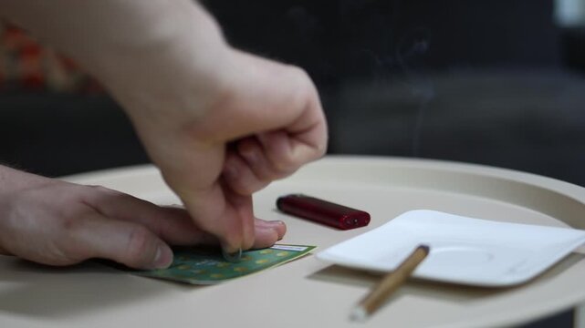 Person scratching a lottery ticket with a coin while smoking a cigarette showing gambling addiction risky behavior and hope for an instant jackpot win