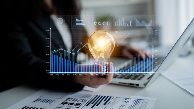 A businesswoman holds a lit light bulb in one hand while working on a laptop surrounded by digital graphs, representing innovative thinking in a corporate environment. Scalp