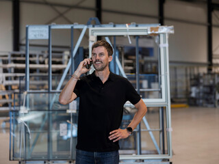 Fototapeta premium Warehouse employee talks on his mobile phone with a hand on his hip, smiling in a storage hall full of storage racks.