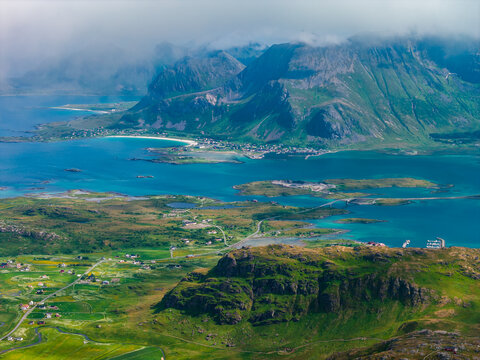 Aerial view over Lofoten Islands, Norway, from the Ryten hike toward sweeping bays and sandbars, glacier blue water, red and white cabins, farms, and low clouds at sunset.