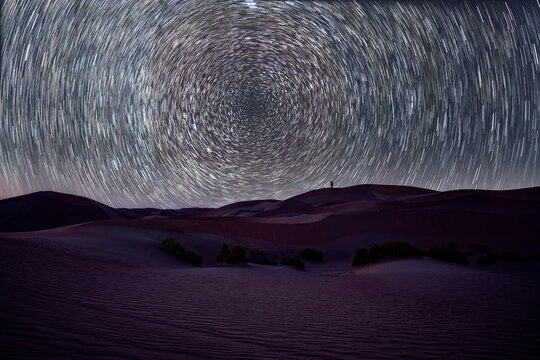 View of a lone person standing on a sand dune under a dramatic circular star trails in the night sky.