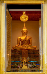Obraz premium Bangkok, with a large, golden Buddha statue inside a temple, its multi-tiered base adorned with Thai patterns. The interior is illuminated with warm light, creating a peaceful atmosphere, with many pe
