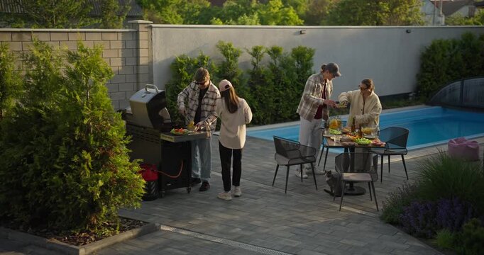 Adults preparing a backyard barbecue beside a swimming pool in a sunny garden
