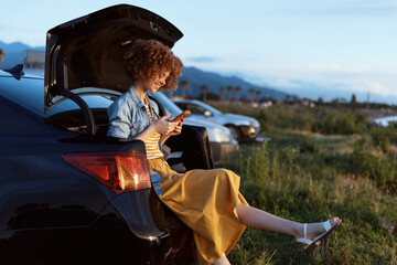 Naklejka premium A woman in a denim jacket and mustard dress sits on a car trunk, using a phone in a sunlit field. A relaxed lifestyle moment outdoors, conveying casual travel and leisure.
