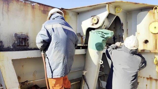 Shipboard routine in action. Crew members work on deck, chipping rust and fixing hatch covers while underway. Honest portrayal of manual labor, teamwork, and seafaring duties.