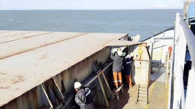 Bulk carrier crew performing maintenance at sea. Sailors use tools to remove rust and repair hatch covers while the ship sails. Authentic industrial maritime work and crew routine.